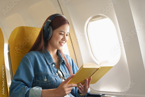 Attractive portrait of an Asian woman sitting in a window seat in economy class reading a book and listening to instrumental music during an airplane flight, travel concept, vacation, relaxation