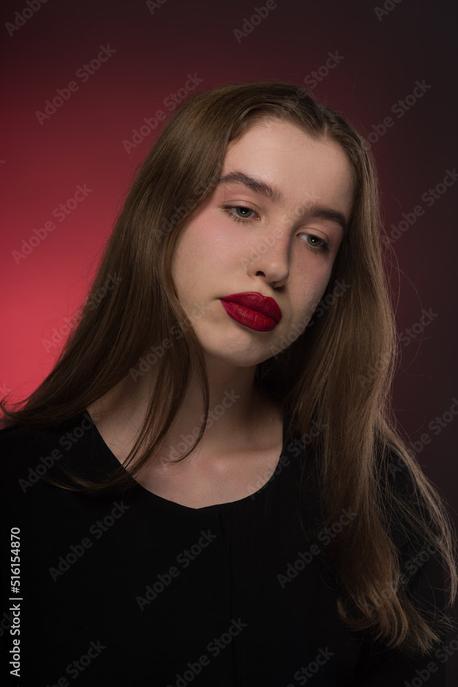 Portrait of a young girl with a dreamy look in a black blouse on a red background. Long brown hair lies loosely on the shoulders. Expressive, plump lips are painted with red lipstick. Close-up. Portrait of a young girl with a dreamy look in a black blouse on a red background. Long brown hair lies loosely on the shoulders. Expressive, plump lips are painted with red lipstick. Close-up.