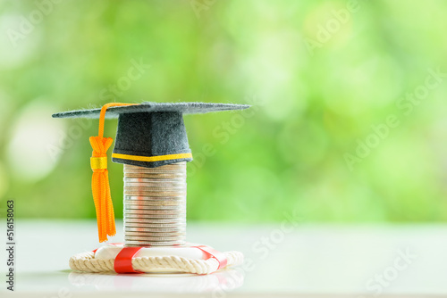 Tuition protection service and tuition refund insurance, financial concept : Black graduation cap or a mortarboard placed higher on top of a coin stack with a red lifebuoy on a table, green background photo