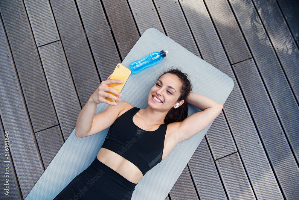 Φωτογραφία Woman lying on yoga mat after exercising, relaxing with smartphone and listening to music