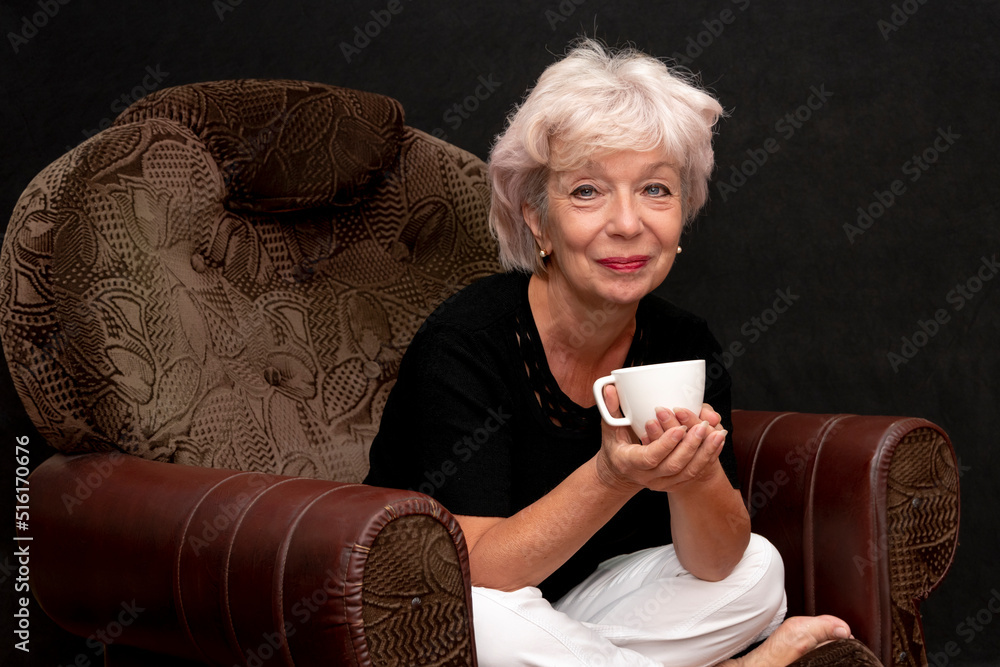 An elderly beautiful blonde woman of 60-65 years old holds a cup in her hand, sits in an armchair on a black background.