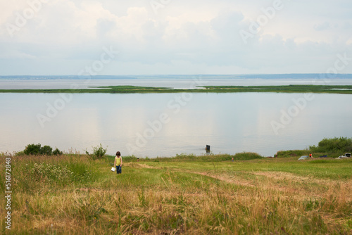 A picturesque bank of a wide river with rare vacationers. Clouds are reflected transparently in the water. On the shore in the grass, a child collects a wild berry.