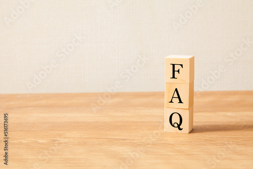 FAQの文字。よくある質問。Frequently Asked Questions。3つの木製ブロックに書かれている。黒い文字。木製テーブルと白い壁紙の背景。左にコピースペース。
