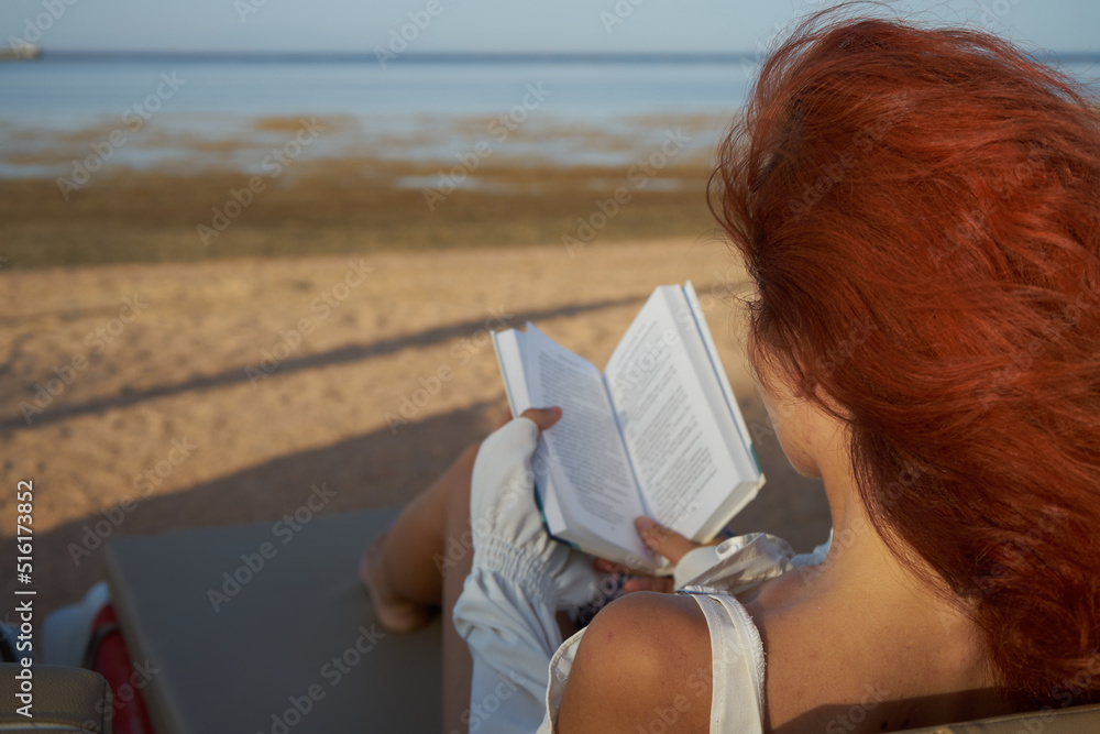 Beauttiful young woman relax at the beach and reading a book. Red hair woman reading book relaxed in deck chair. Summer holiday vacation. Beauttiful young woman relax at the beach and reading a book. Red hair woman reading book relaxed in deck chair. Summer holiday vacation.