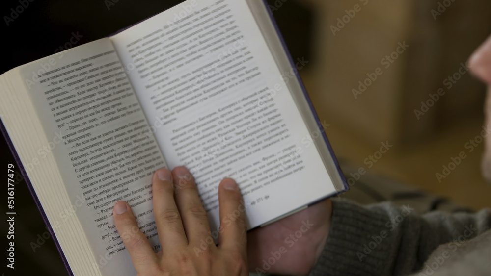 Naklejka premium Over the shoulder view of a man reading a book at home. Video. Middle aged male sitting and reading a paper book, turning pages, relaxing at home.