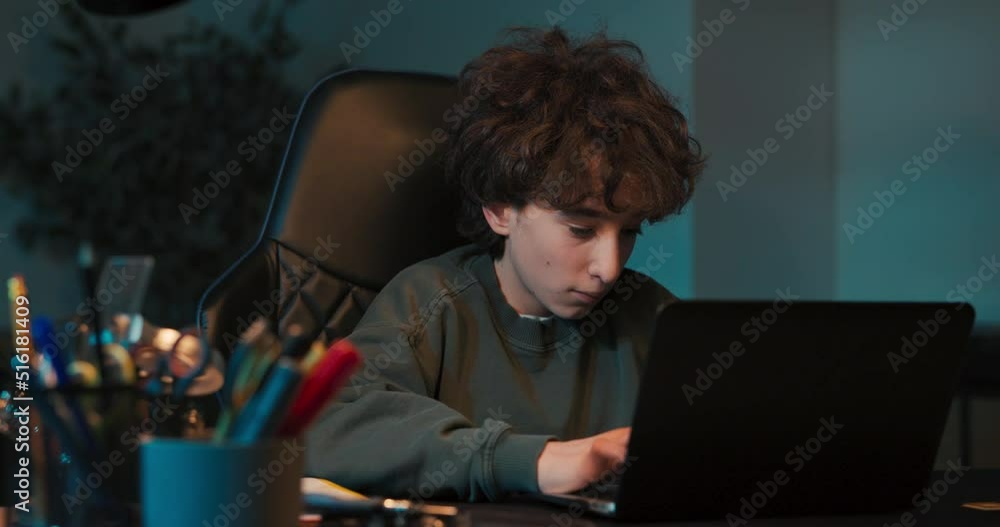 Teenage boy doing homework using computer sitting by desk in room alone. Teenager watching video and browsing internet on laptop. Teenage boy doing homework using computer sitting by desk in room alone. Teenager watching video and browsing internet on laptop.