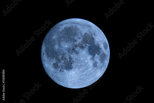 Blue full moon on dark black sky background, shot from surface of earth with very long telephoto lens. Close-up of moon glowing at clear sky.