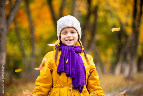 Portrait of smiling little adorable girl in autumn park with yellow and green trees and flying leaves