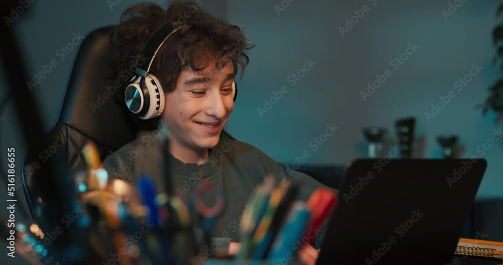 A young boy with curly hair is happy with remote learning. A student sits in front of a computer with wireless headphones listening to a teacher lecture. The lessons take place over the Internet. A young boy with curly hair is happy with remote learning. A student sits in front of a computer with wireless headphones listening to a teacher lecture. The lessons take place over the Internet.