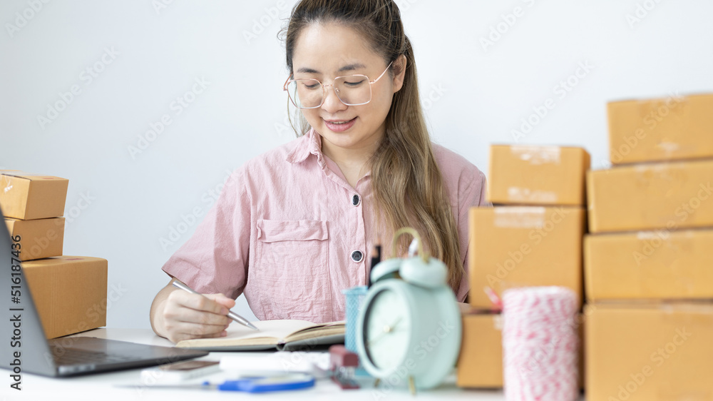 Young Asian woman is writing down the customer's details and addresses on the notebook or box in order to prepare for shipping according to the information, New kind of business for young, Sell online Young Asian woman is writing down the customer's details and addresses on the notebook or box in order to prepare for shipping according to the information, New kind of business for young, Sell online