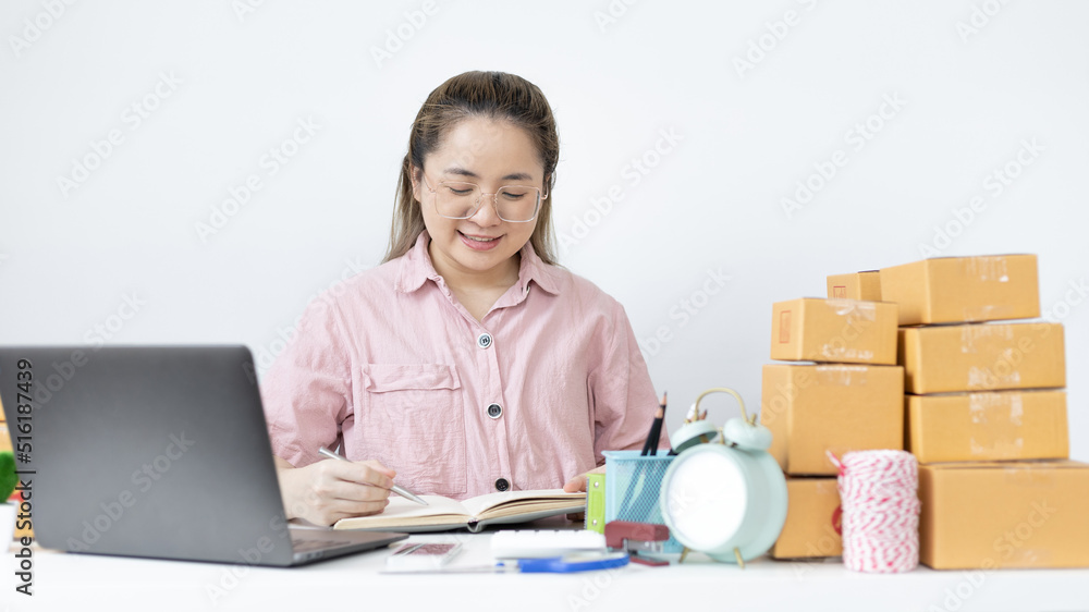 Young Asian woman is writing down the customer's details and addresses on the notebook or box in order to prepare for shipping according to the information, New kind of business for young, Sell online Young Asian woman is writing down the customer's details and addresses on the notebook or box in order to prepare for shipping according to the information, New kind of business for young, Sell online