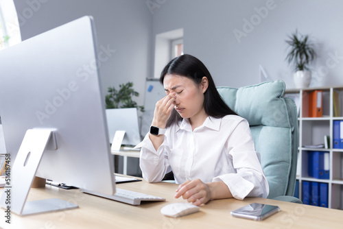 Exhaustion at work. Tired young Asian woman, businesswoman, manager, accountant, freelancer sits in a chair in the office at the desk, works at the computer, needs a break.