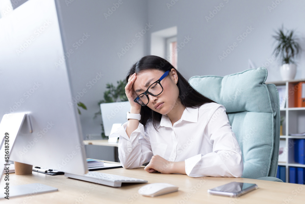 Exhaustion at work. A young Asian woman in glasses holds her head, feels pain, grimaces. Sitting in the office at the desk, working at the computer, exhausted. Businesswoman, accountant, freelancer. Exhaustion at work. A young Asian woman in glasses holds her head, feels pain, grimaces. Sitting in the office at the desk, working at the computer, exhausted. Businesswoman, accountant, freelancer.