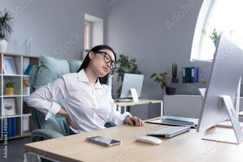 Young beautiful azitaka businesswoman wearing glasses feeling exhausted at work. Holds his back with two hands, massages, feels pain and fatigue. Sitting at the desk at the computer, needs a break.