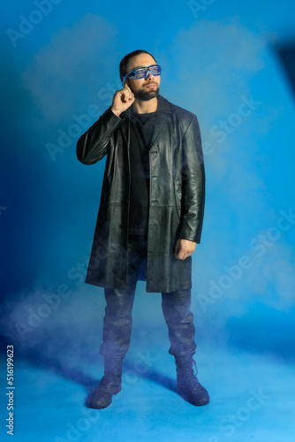 portrait of a man in a studio in a black leather coat. model with a beard on a blue background. trendy luminous glasses on the face