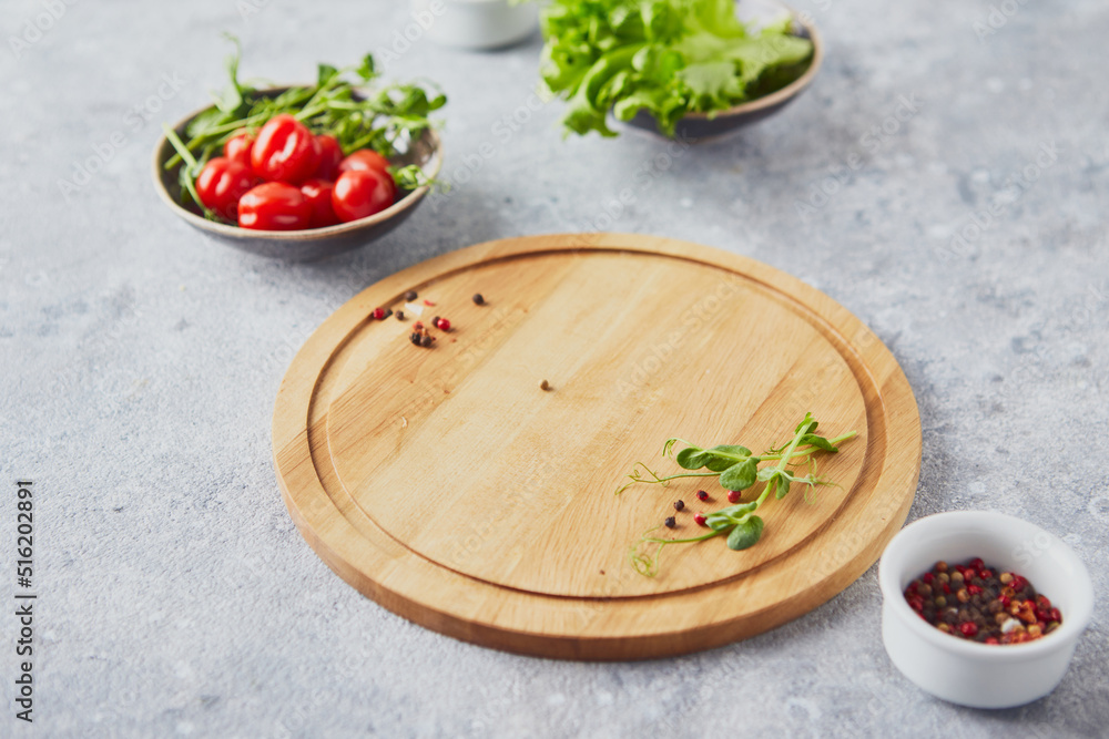 Wooden round cutting board, greens, vegetables and seasonings on light background Wooden round cutting board, greens, vegetables and seasonings on light background