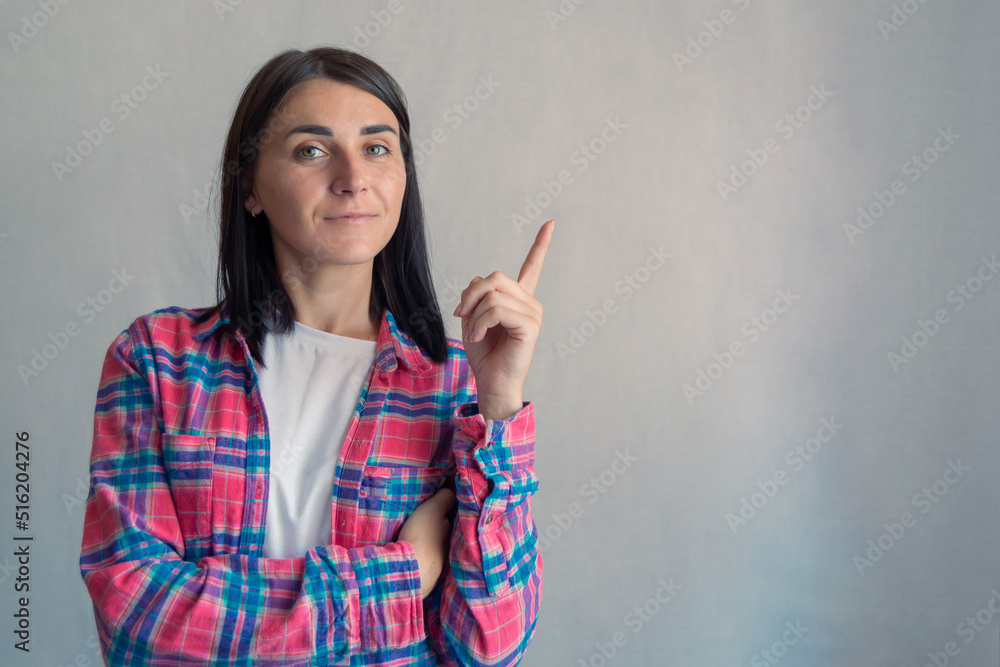 Young attractive confident woman in casual shirt showing pointing finger at something or copy space empty text offer area. Grey color background. Portrait of Brunette pretty girl Young attractive confident woman in casual shirt showing pointing finger at something or copy space empty text offer area. Grey color background. Portrait of Brunette pretty girl