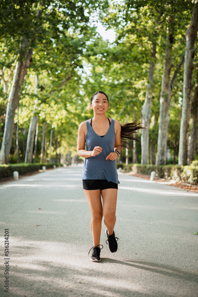 Happy asian sport girl jogging in the park surrounded by green trees