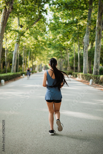 Vertical back shot of athlete girl jogging in the park surrounded by green trees. © Pablo Rasero