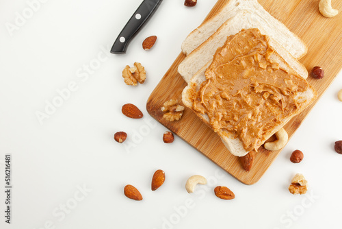 Square bread for toast with peanut butter on a wooden board. Nuts, a knife and a wooden cutting board with a sandwich and slices of bread on a white table.