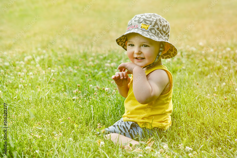 Happy toddler baby boy sit on green grass in nature. Smiling child in shorts, yellow t-shirt and hat plays in the summer in the park. Kid age one year Happy toddler baby boy sit on green grass in nature. Smiling child in shorts, yellow t-shirt and hat plays in the summer in the park. Kid age one year