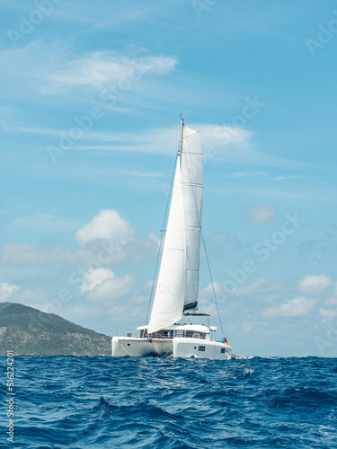 Luxurious white catamaran is sailing at full sail on the blue ocean water, moving away from the island on a sunny summer day. The concept of yachting, sea travel