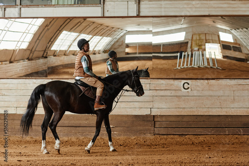 Full length side view at young woman riding horse in indoor arena or practice stadium with mirrors, copy space