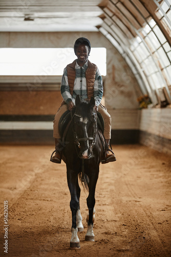 Vertical portrait of smiling young woman riding horse in indoor arena at horse ranch or practice stadium
