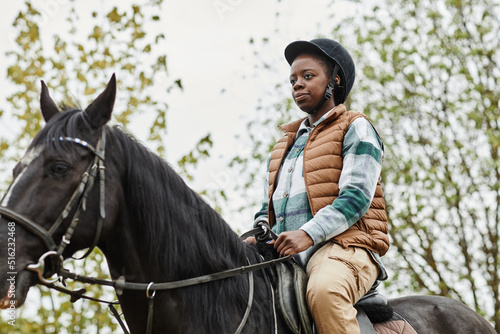 Portrait of young black woman riding horse outdoors and wearing protective helmet, copy space