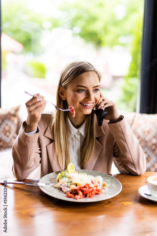 Young smiling happy woman sit alone at table in cafe shop eat breakfast talk speak on mobile cell phone drink coffee relaxing in restaurant during free time indoors.