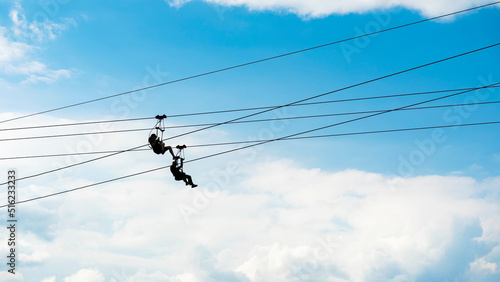 Zipline riders, silhouettes of two people on blue sky background