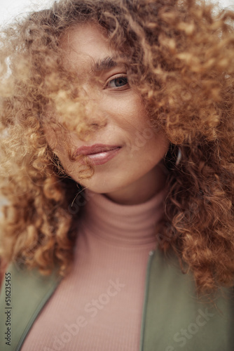 Young stylish woman in beige turtleneck and casual jacket with her long and thick wavy hair covering part of face looking at camera