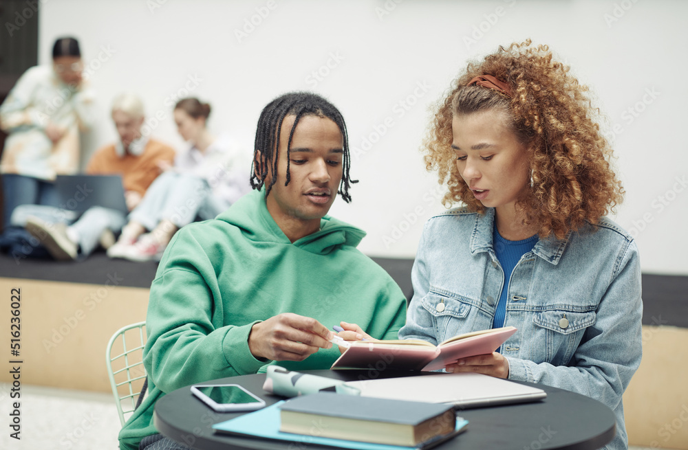 Teenage guy and his girlfriend discussing lecture notes in copybook before lesson or seminar while sitting by table against their classmates Teenage guy and his girlfriend discussing lecture notes in copybook before lesson or seminar while sitting by table against their classmates