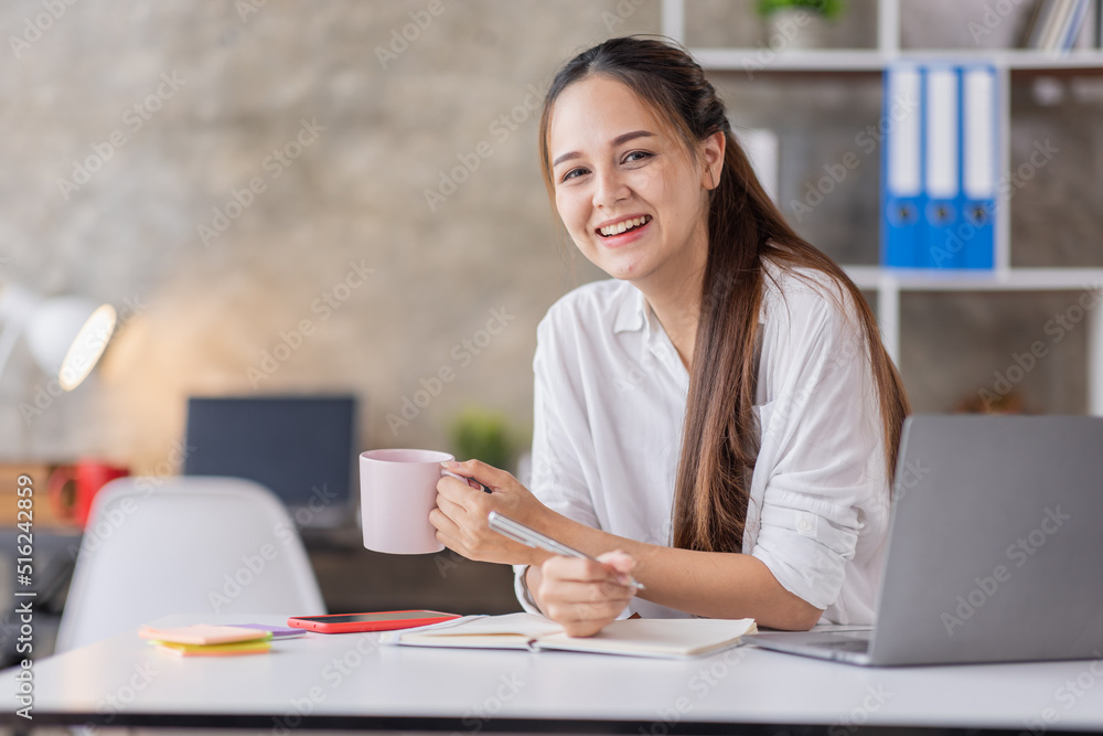 Beautiful young Asian woman working on a laptop computer while sitting in the office room, drinking coffee Beautiful young Asian woman working on a laptop computer while sitting in the office room, drinking coffee