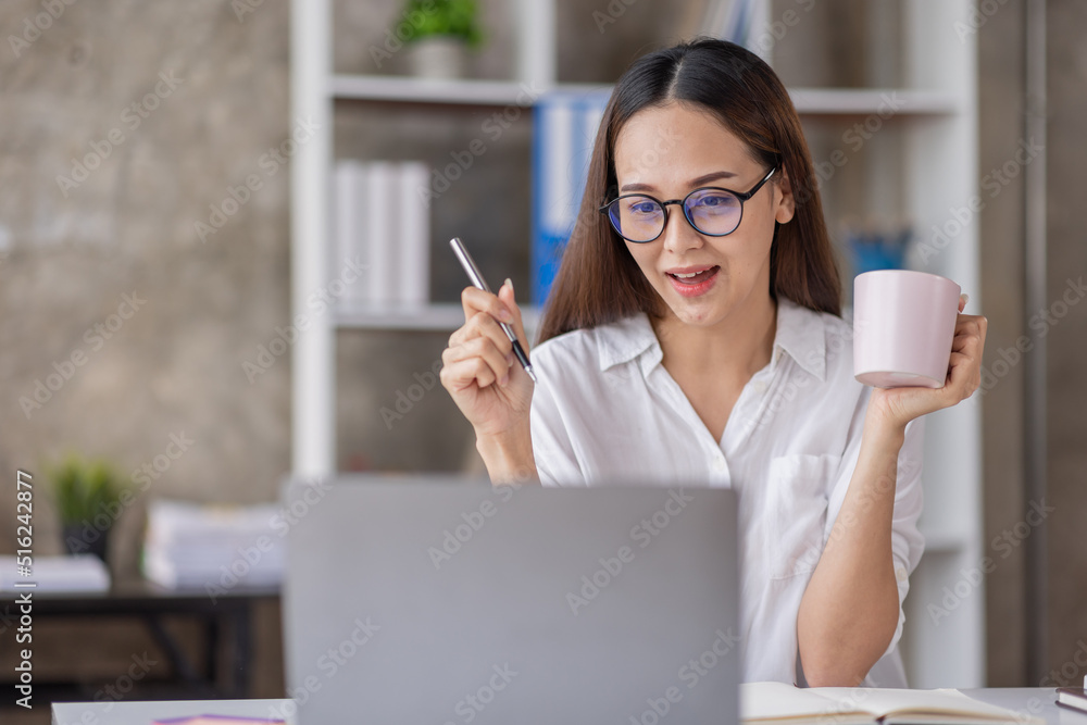 Beautiful young Asian woman working on a laptop computer while sitting in the office room, drinking coffee Beautiful young Asian woman working on a laptop computer while sitting in the office room, drinking coffee