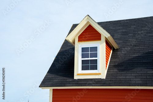 The exterior of a red wooden clapboard building with a steep black shingled roof. A closed glass window and dormer with white trim are in the attic of the house. The background is a cloudy blue sky.