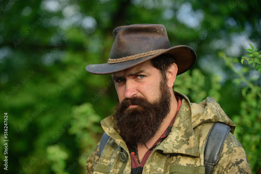 Portrait of adventure serious man extreme explorer. Man hunter in camouflage outdoor. Brutal hunter, bearded man in the wild forest in the autumn. Portrait man in cowboy hat.