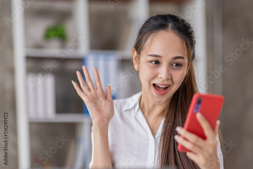 Happy Asian young woman waving hands looking at web camera using phone for video call at home, smiling business woman having fun greeting online by phone webcam making video call via application