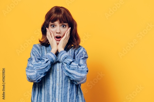 a surprised, sweet woman stands on a yellow background in a blue shirt and holds her hands near her face. Horizontal photo with empty space