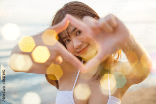 Close up Asian happy young woman looking through making shape camera frame made from her fingers on the beach at sunset, Composition finger frame