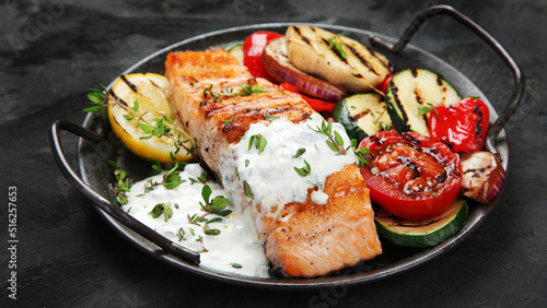 Salmon steak with vegetables and white sauce on dark background.