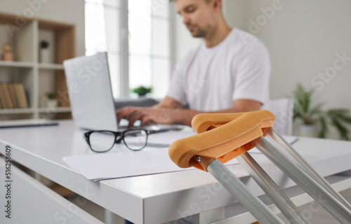 Young businessman has injury and uses crutches. Close up of crutches on background of concentrated man working on laptop in office. Medical equipment and business concept. Selective focus. photo