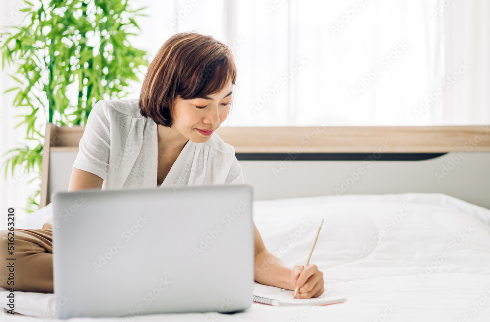 Young smiling happy beautiful asian woman relaxing using laptop computer in the bedroom at home.Young creative girl working and typing on keyboard.work from home concept Young smiling happy beautiful asian woman relaxing using laptop computer in the bedroom at home.Young creative girl working and typing on keyboard.work from home concept