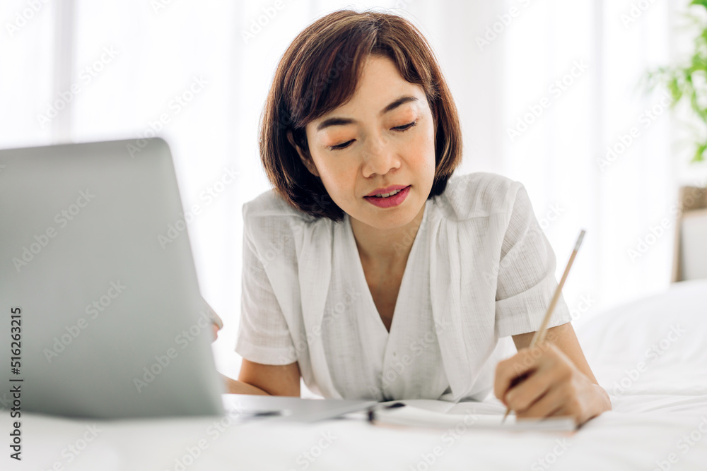 Young smiling happy beautiful asian woman relaxing using laptop computer in the bedroom at home.Young creative girl working and typing on keyboard.work from home concept Young smiling happy beautiful asian woman relaxing using laptop computer in the bedroom at home.Young creative girl working and typing on keyboard.work from home concept