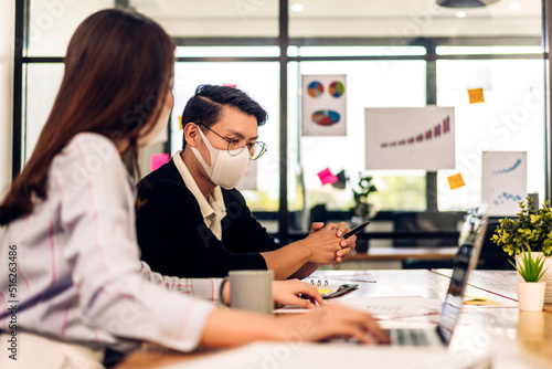 Asian business man and woman using laptop computer working and planning meeting in quarantine for coronavirus wearing protective mask with social distancing while sitting on office desk