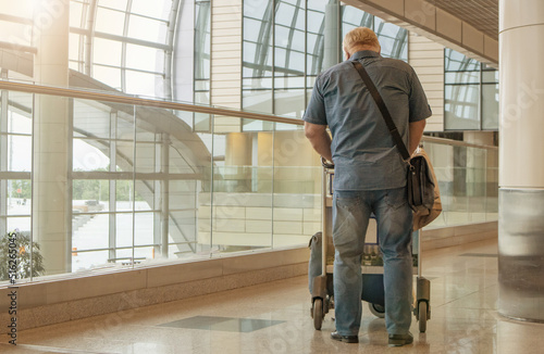 A full-length figure of a man in blue jeans and a shirt, with a luggage cart, against the background of the airport interior, a copy of the space on the left