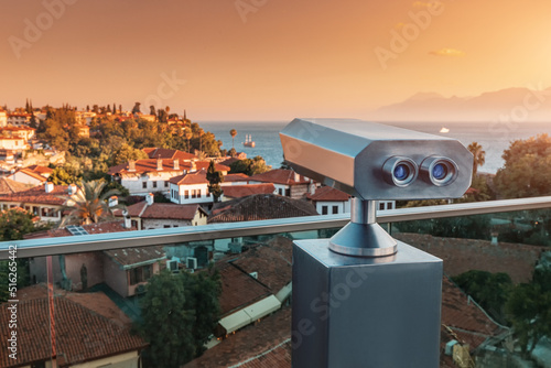 Tourist optical telescope on the observation deck viewpoint overlooking the old resort town with orange roofs during a colorful sunset in Antalya city