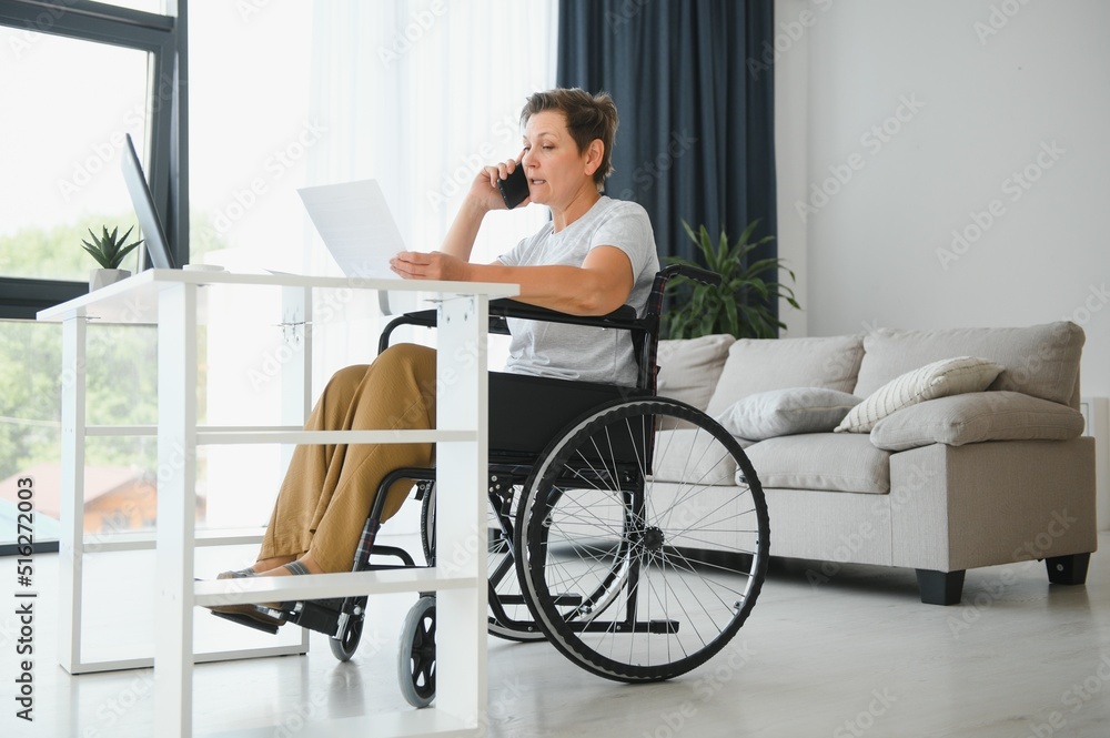 Freelancer in wheelchair using laptop near notebook and papers on table Freelancer in wheelchair using laptop near notebook and papers on table