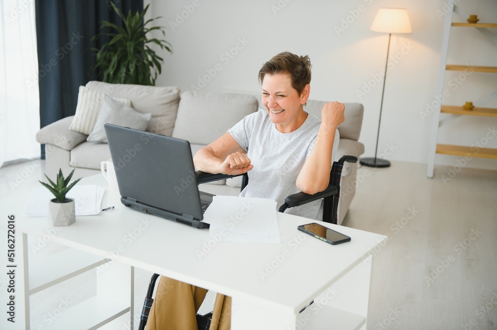 Freelancer in wheelchair using laptop near notebook and papers on table