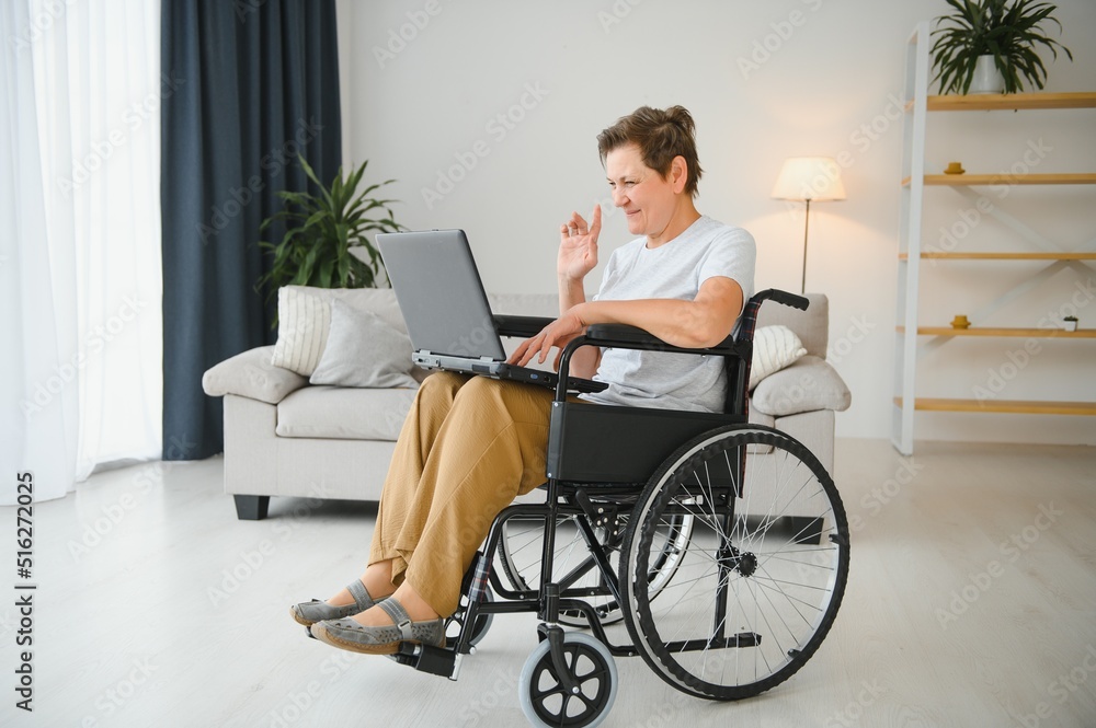 Middle age grey-haired disabled woman having video call sitting on wheelchair at home. Middle age grey-haired disabled woman having video call sitting on wheelchair at home.